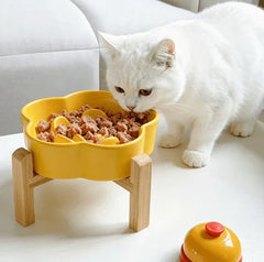 White cat eating from a Petroom yellow flower-shaped ceramic slow feeder bowl on a wooden stand.