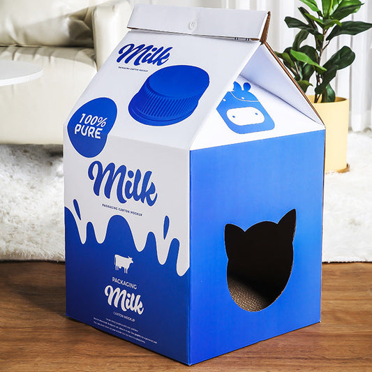 A milk carton-shaped cat tree or playhouse made from blue and white cardboard, placed on a wooden floor in a living room setting with a sofa and plant in the background.