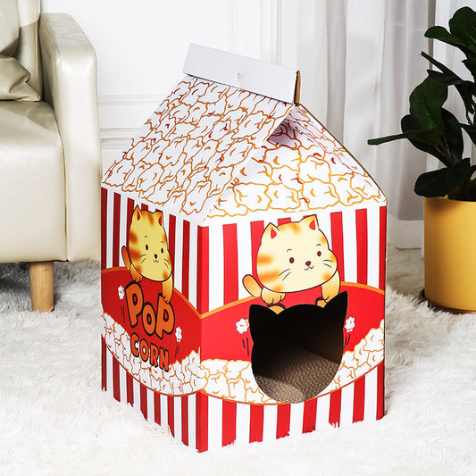 A cardboard cat house designed like a popcorn bag, featuring red and white vertical stripes, a white top with orange popcorn graphics, cartoon cat illustrations, and a black cat hole in the front, placed on a white rug in a living room setting.