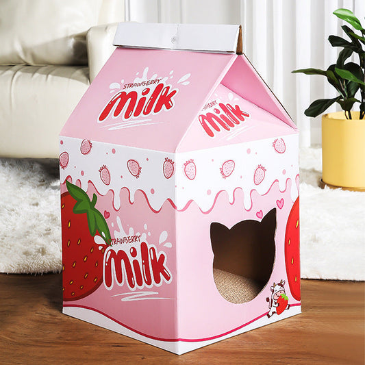 A pink cardboard cat house designed to look like a milk carton, featuring strawberry illustrations and a cat-shaped window cutout, set on a wooden floor in a living room.