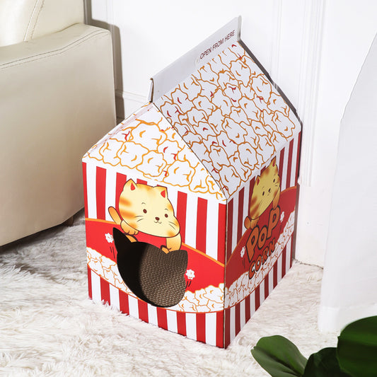 A house-shaped cardboard cat playhouse designed to look like a popcorn box, with red and white vertical stripes, a cartoon cat illustration, and a black mesh window, placed on a white rug next to a sofa.