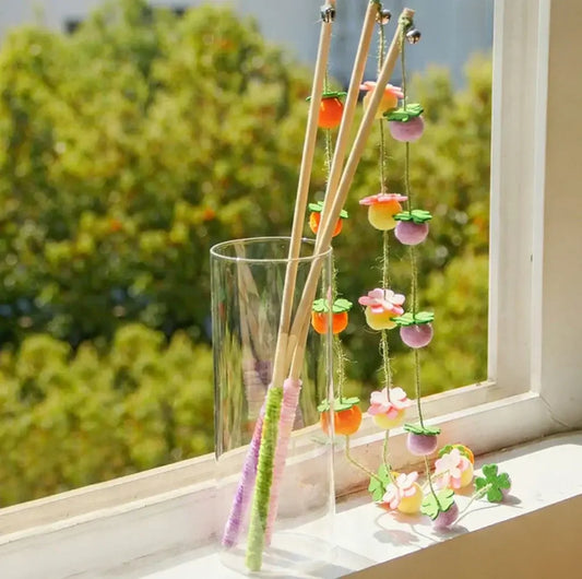 Colorful cat wand toys with felt flowers and bells, displayed in a glass vase by a sunny window.